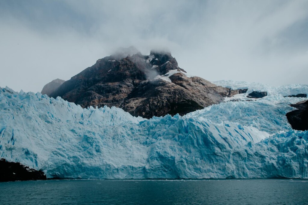 A bright blue glacier sits in front of a gray mountain on a cloudy day