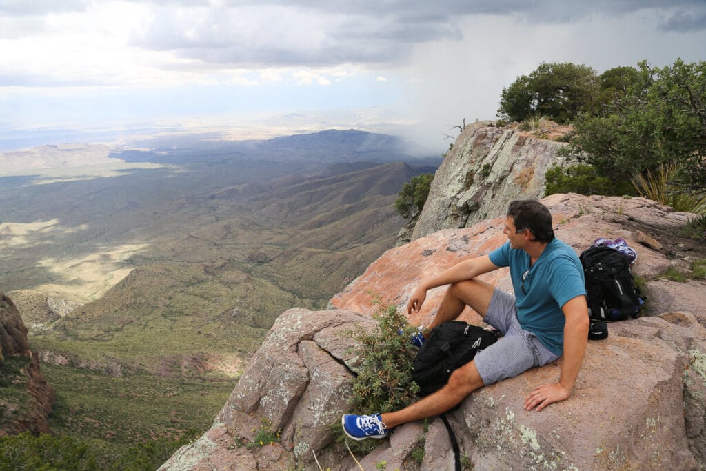 A man sits on a rocky cliff in a blue shirt looking into the distance