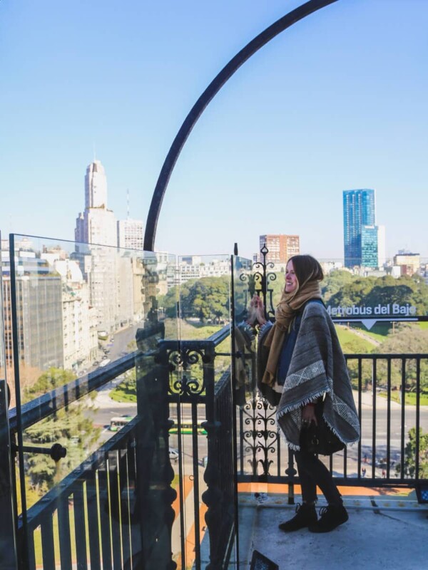 A woman with a poncho and beige scarf stands on a roof terrace looking at the city