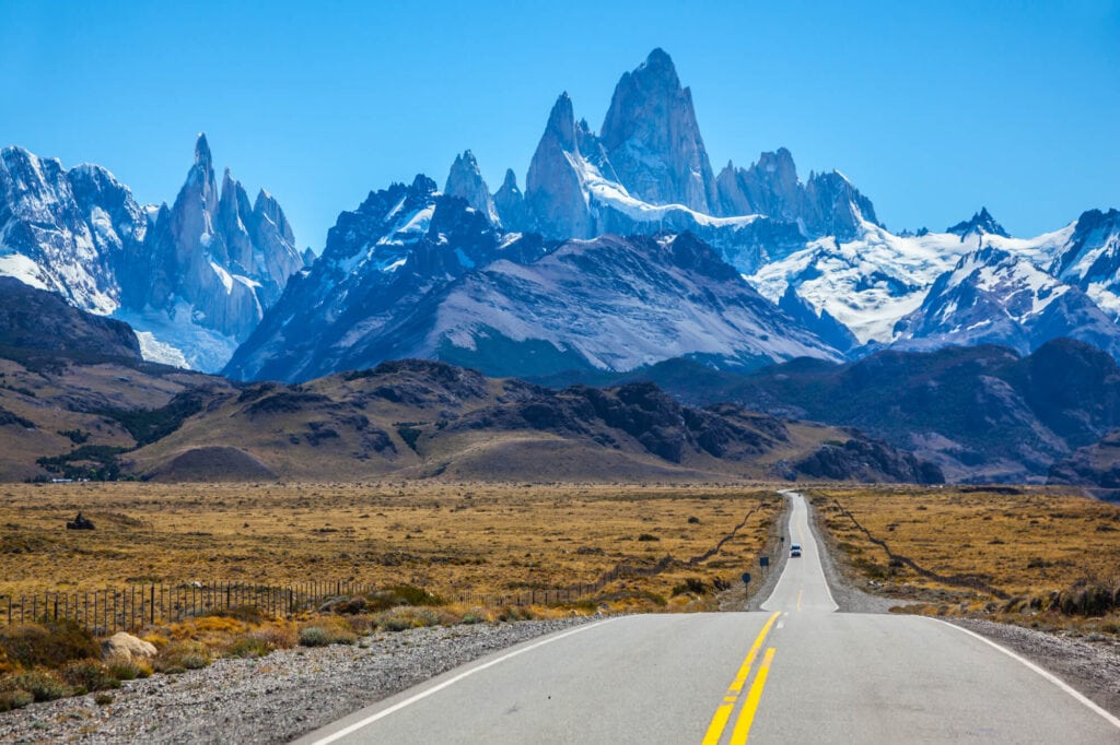 A highway leads from the photographer to a mountain range of rocky peaks and a bright blue sky.