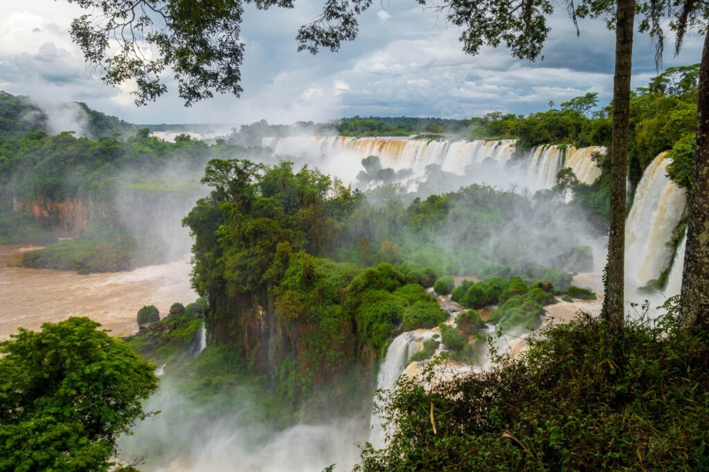 A line of waterfalls of brown water pour down in a green jungle with mist rising up from the river below
