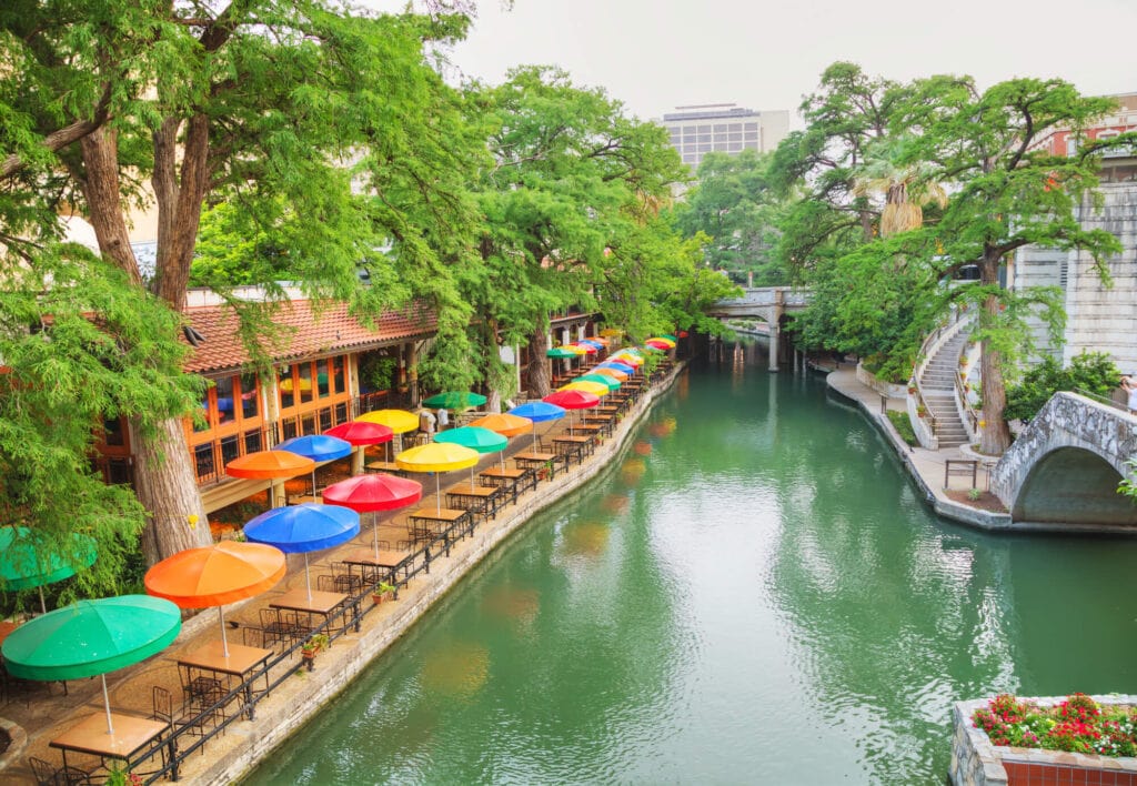 A green river with a sidewalk on both sides and a rainbow of umbrellas over tables of a restaurant on the shore