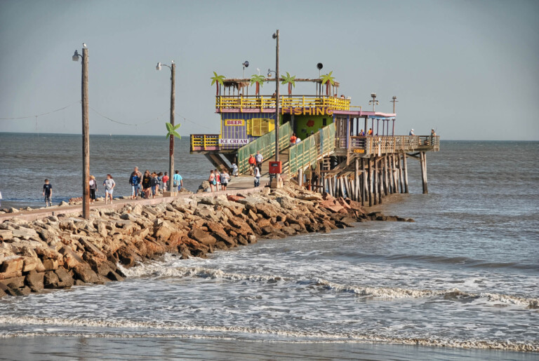 A rocky pier juts out over a brown ocean with a colorful restaurant at the end over the water