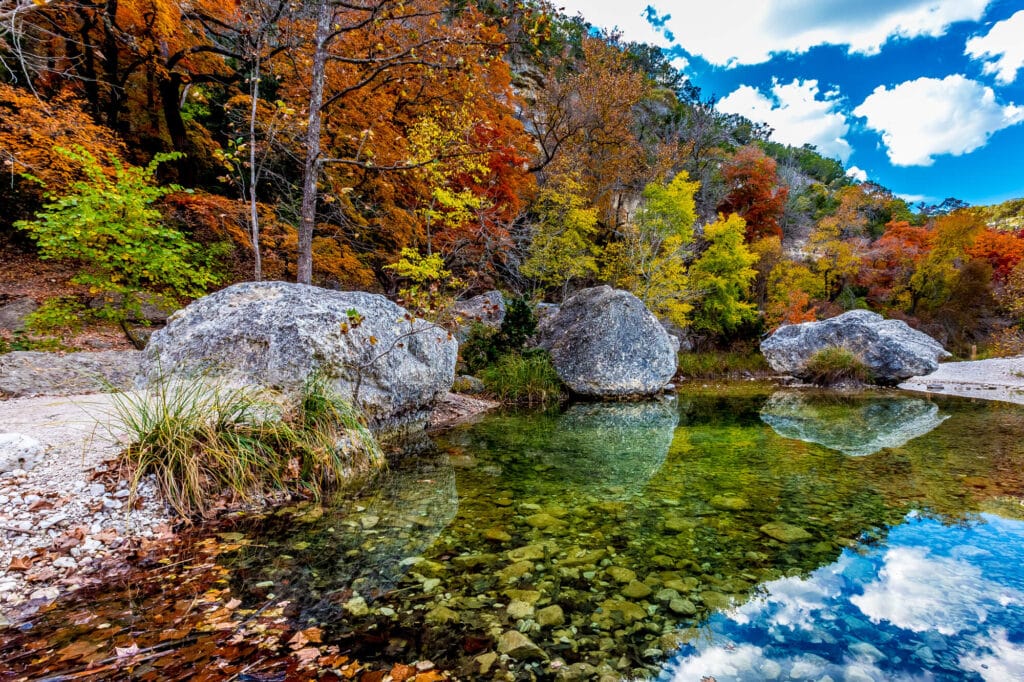 A transparent lake shows the rocks at the bottom with boulders on the shore and orange fall foliage behind them