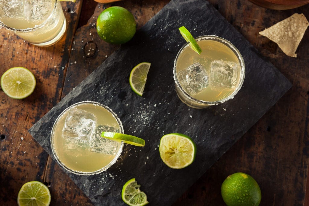 Two margaritas on the rocks on a stone serving platter surrounded by limes