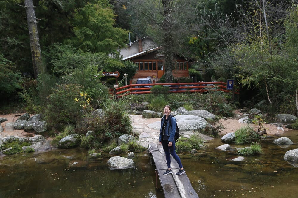 A woman stands on a wood plank bridge over a creek in a pine forest in front of a wooden cabin