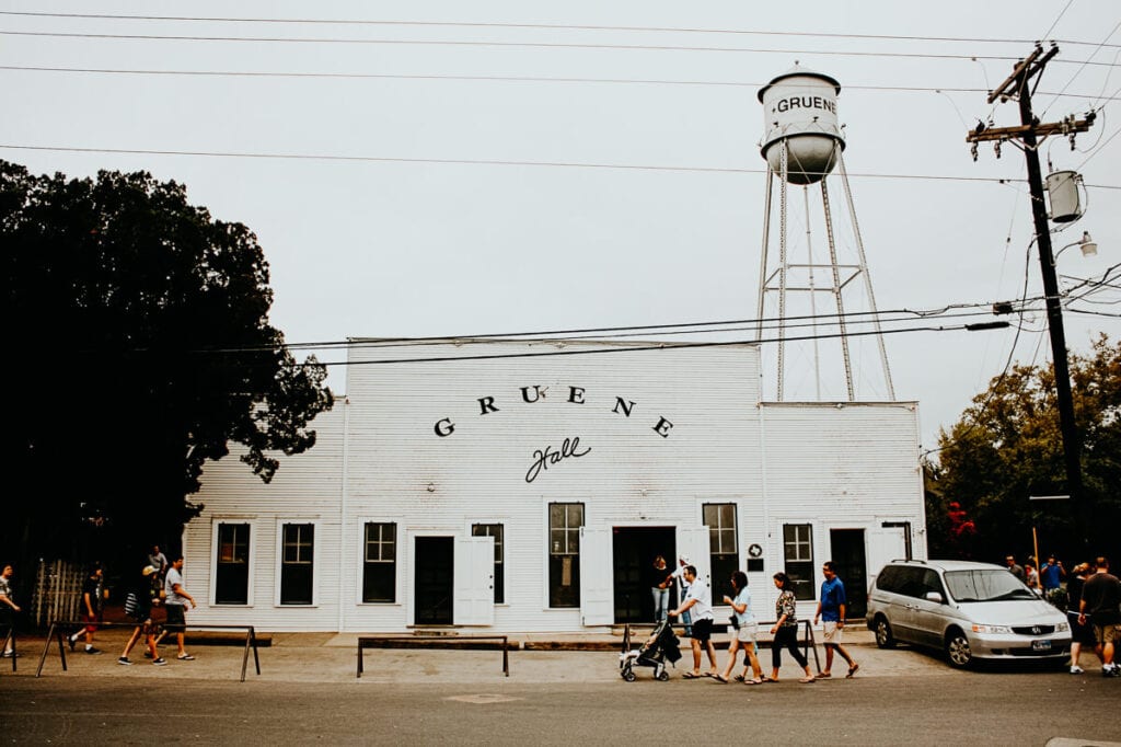 Four people walk in front of a white wooden building with a large metal water tank overhead