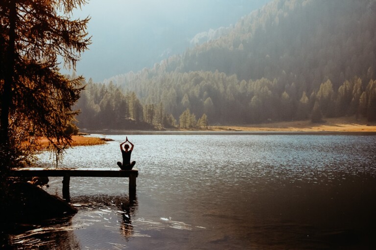 A woman sits on a wooden pier doing yoga by a forest
