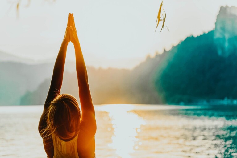 A woman is doing yoga with her hands together overhead with the sun bathing her in orange light in front of a lake