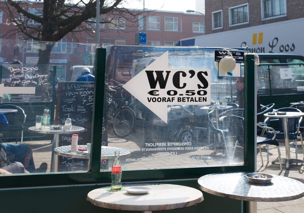 Two round tables at a sidewalk cafe by a glass divider with a WC sign