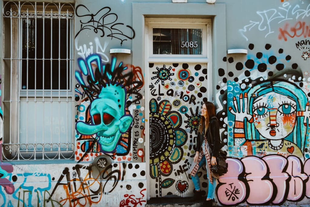 A woman in a black leather coat stands in front of a graffiti covered wall leaning on a door frame