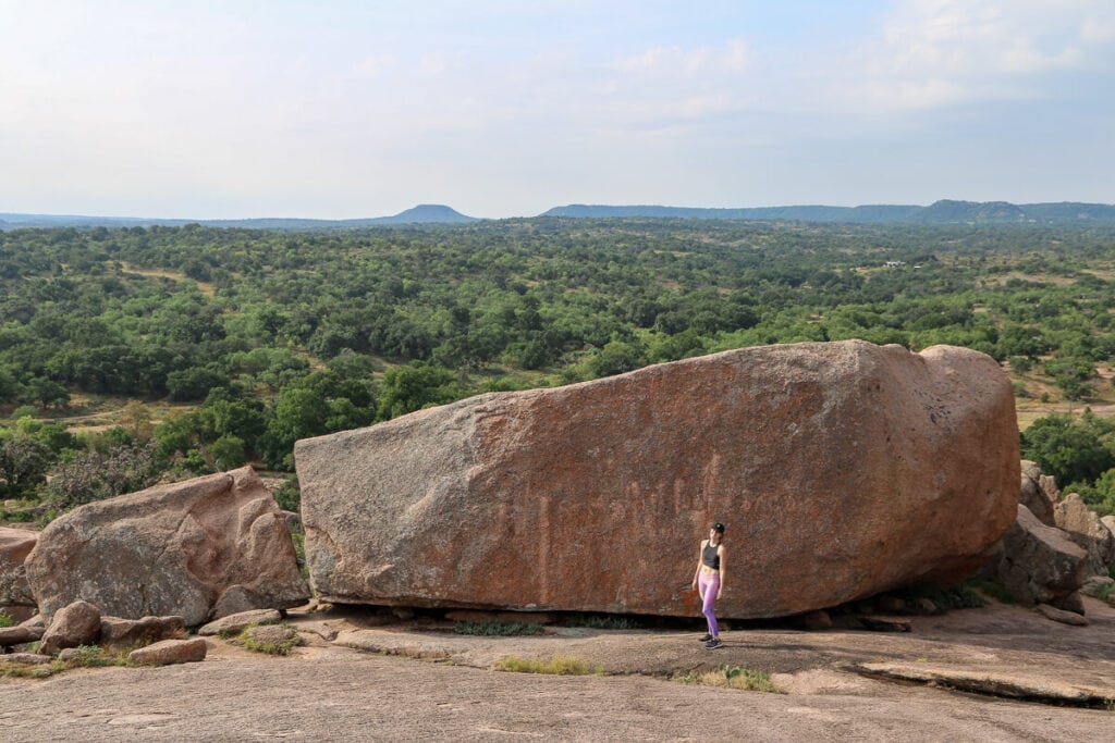 A woman is standing in front of an enormous granite boulder with rolling green hills in the background