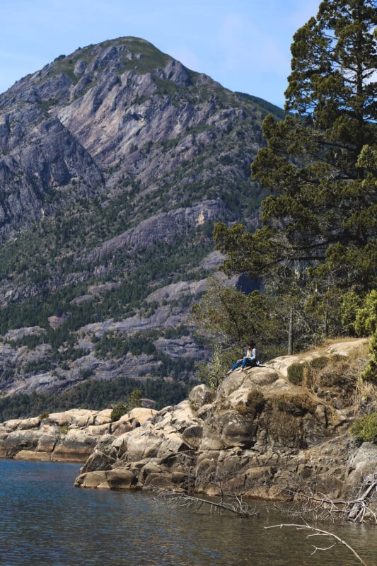 A woman sits on a boulder over a lake with a pine tree and mountain in the background