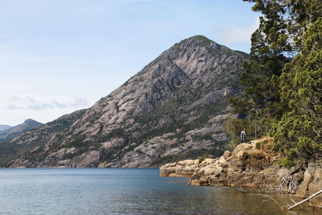 A woman sits on a boulder over a lake with a pine tree and mountain in the background