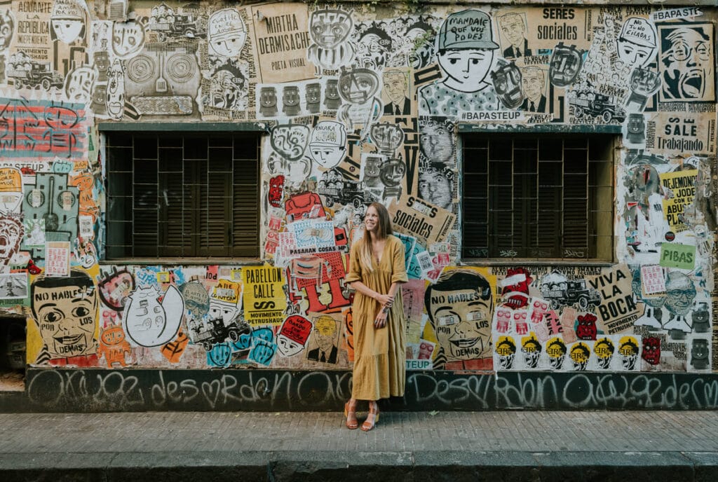 A woman in a beige dress leans against a graffiti covered wall