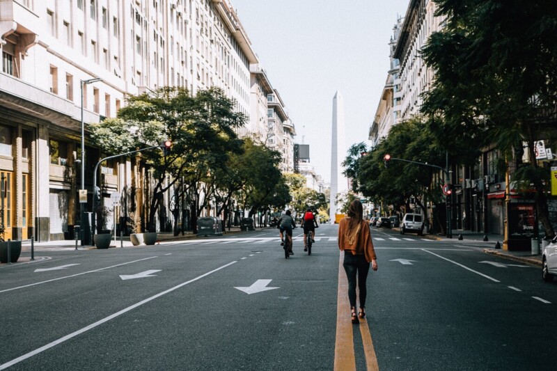 A woman walks away from the camera down the middle of a street towards an obelisk