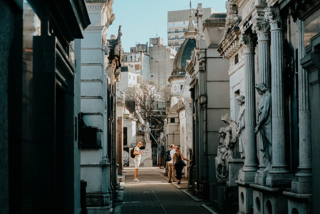 3 people stand in a passageway between above ground mausoleums and crypts.