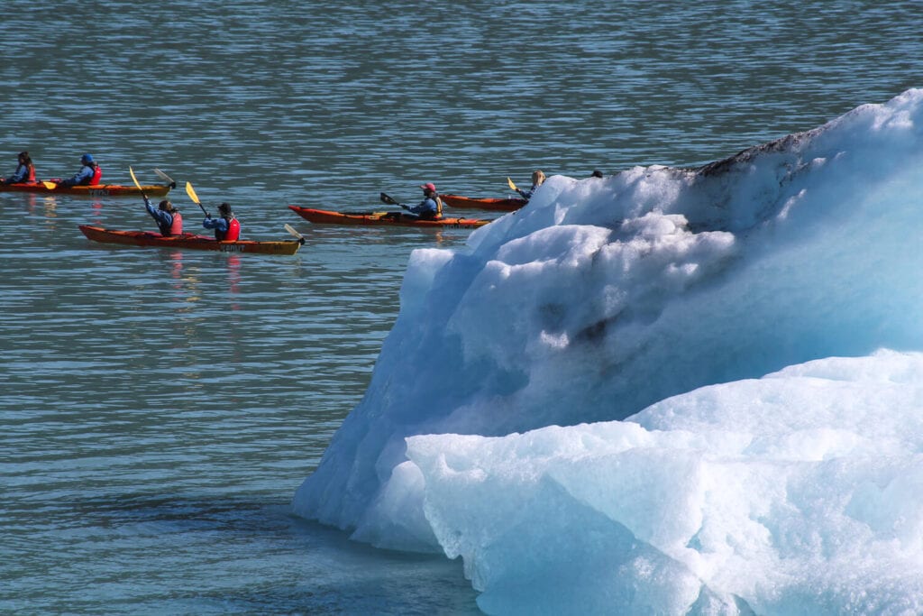 Four red kayaks paddle by an enormous iceberg.