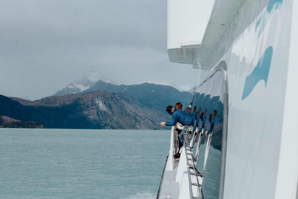 A man in a blue coat leans against the rail of a catamaran watching the mountains in the distance.