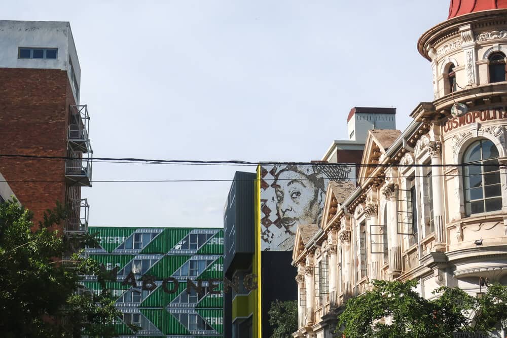 A historic colonial building stands next to a modern building of shipping containers