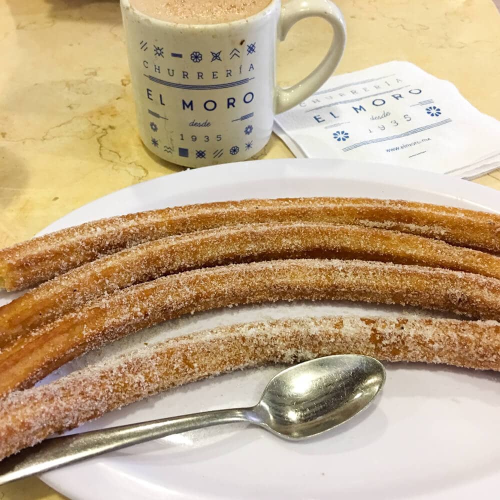 Four churros on a plate next to a cup of hot chocolate