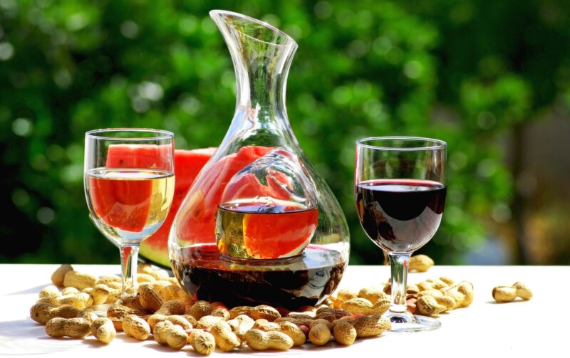 A carafe of red wine by two wine glasses on a table with peanuts and watermelon