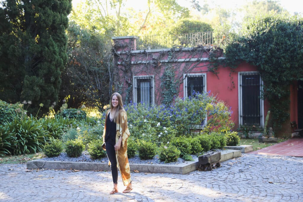 A woman stands on cobblestone in front of a garden and colonial style pink home