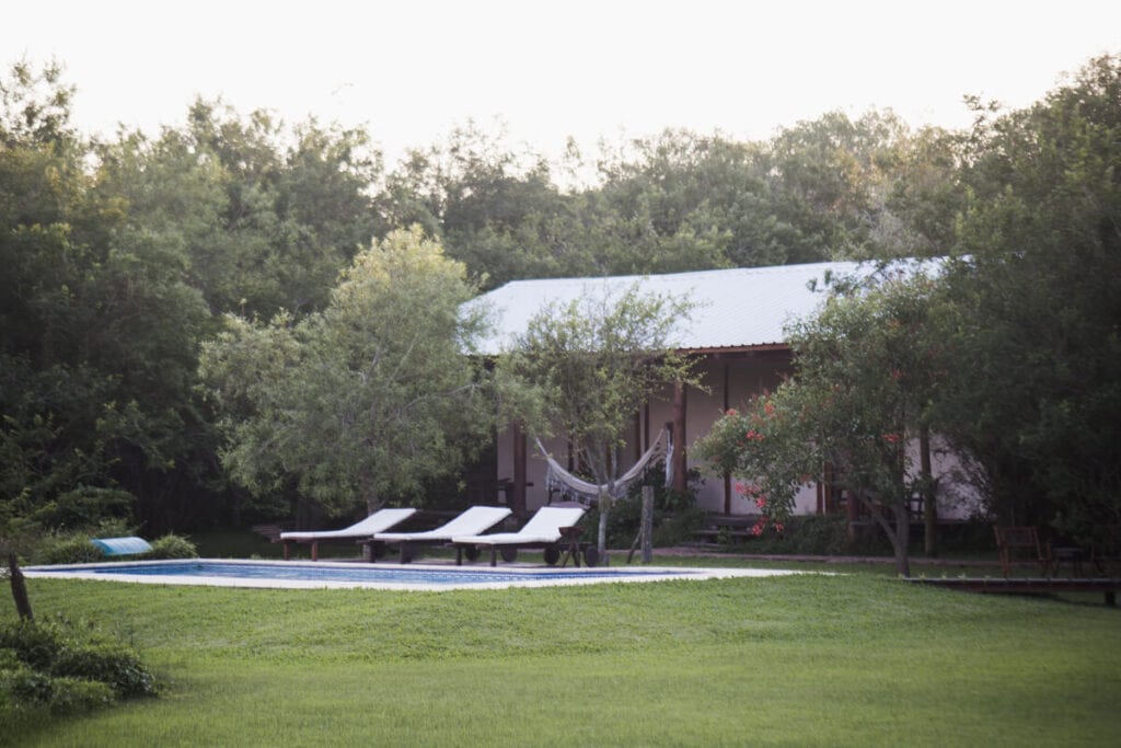 A single story building with a tin roof behind a pool and three lounge chairs