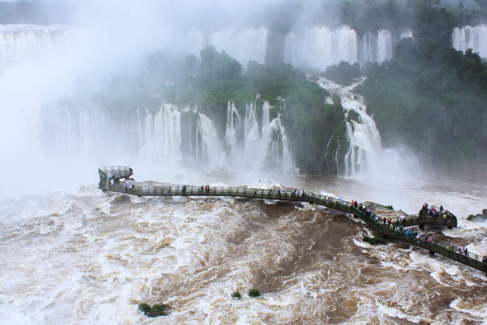 A metal catwalk over a brown river in front of waterfalls