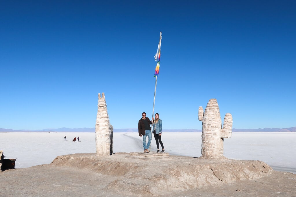 A man and woman stand under a flat pole next to salt statues of a llama and a cactus