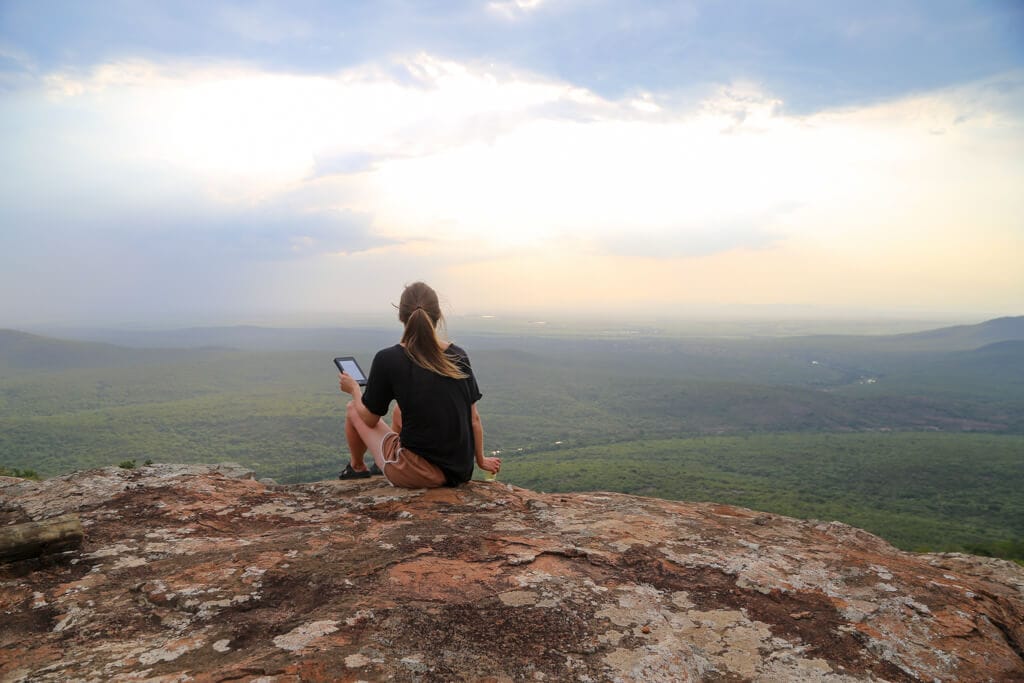 A woman in a black t-shirt sits on a cliff's edge at sunset