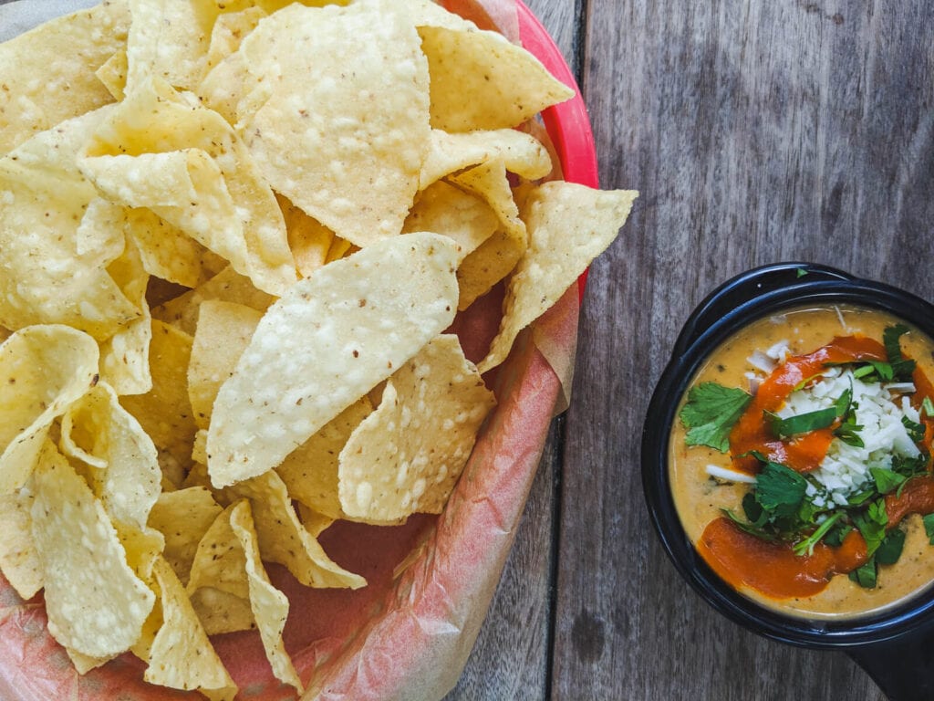 A basket of chips on a table next to a cup of queso dip in texas