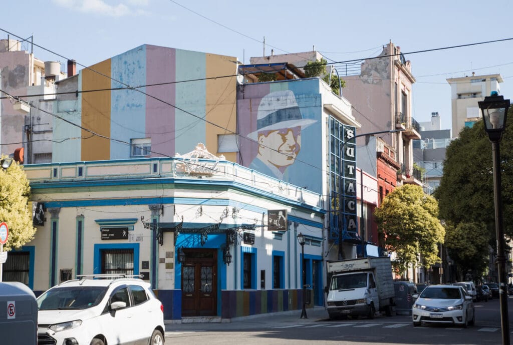 Cars drive by a street corner with a carlos gardel mural painted on the upper floors