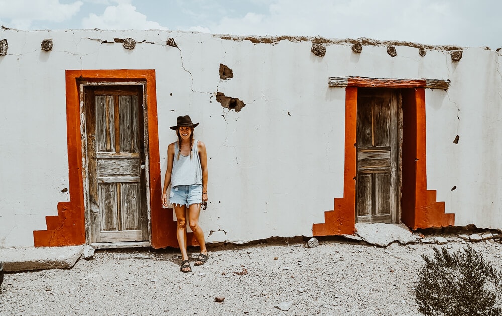 A woman leans against a red and white stucco building in the desert