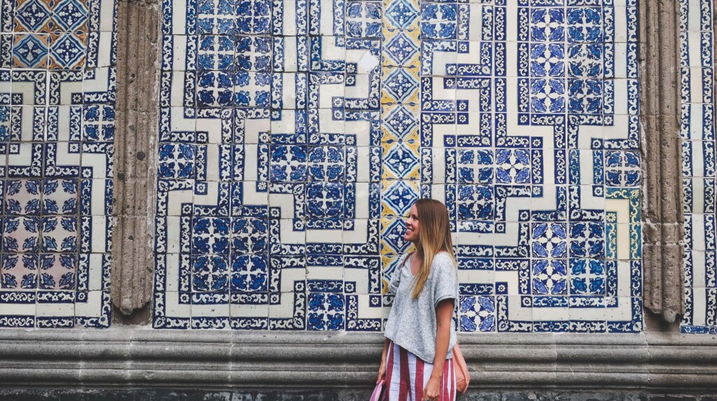 A woman in a grey sweater stands in front of a blue and white tiled wall