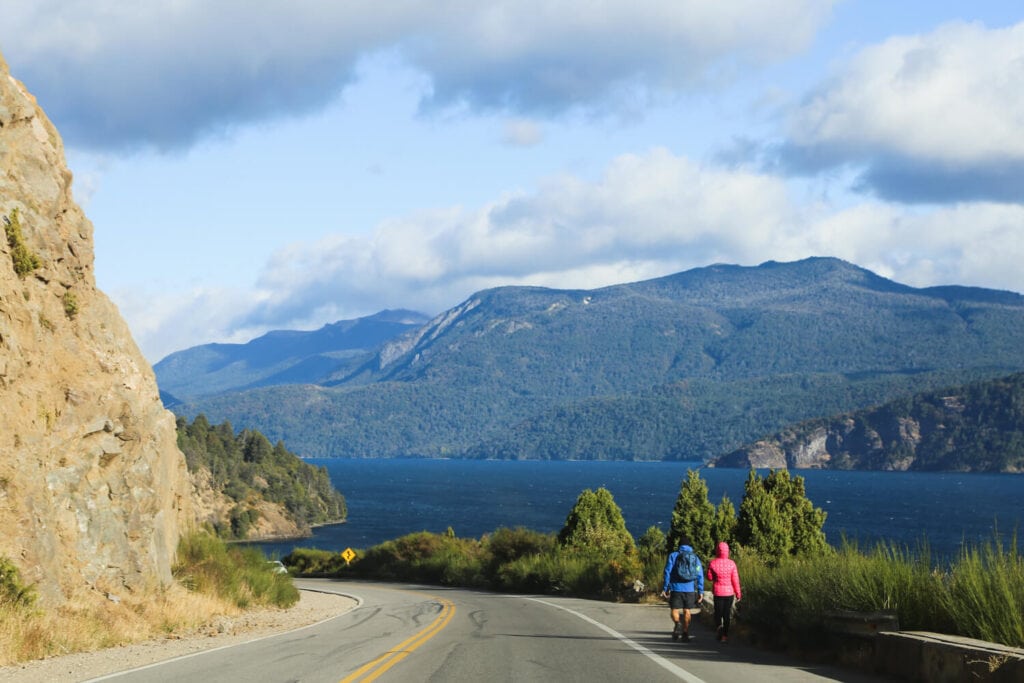 Two people walk beside a highway next to a lake