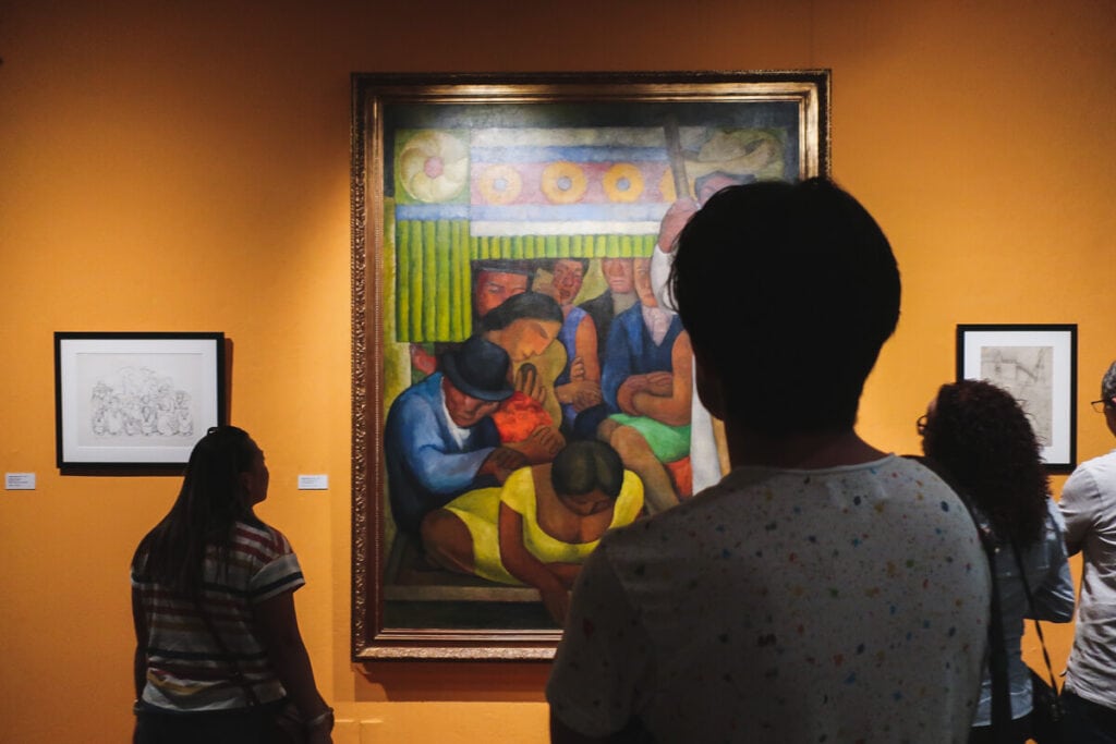 Three people stand in front of a painting on an orange wall in a museum