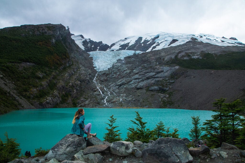 A woman sits on a rock in front of a turquoise glacier lake