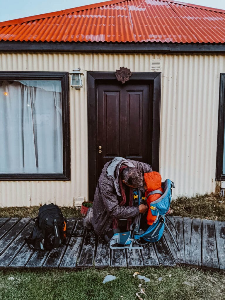 A man straps a baby into his backpack in front of a cabin