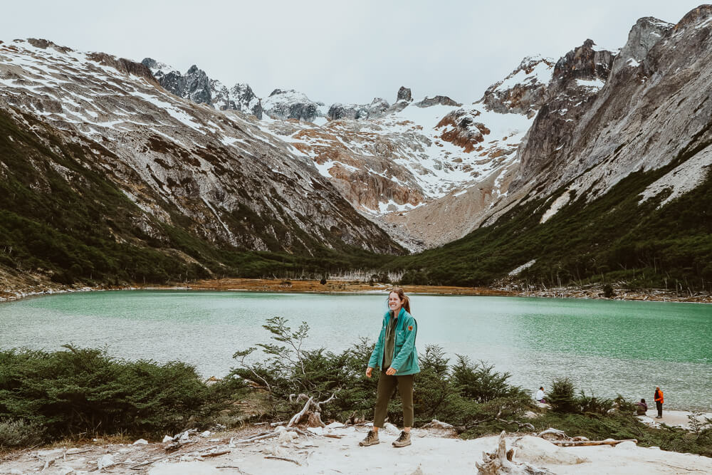 A woman stands in front of a turquoise lake in the mountains