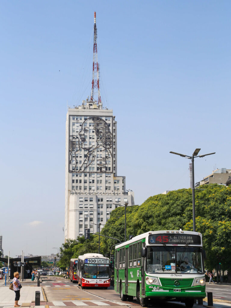 Red and green city buses drive down a wide avenue in front of a skyscraper with an Evita sign on it