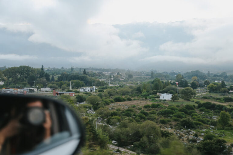 A woman takes a photo of a lush green valley and appears in a side view mirror