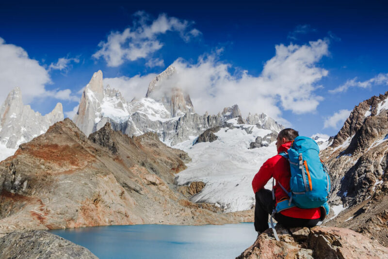 A man in a red sweater and blue backpack sits on a rock at Laguna de los Tres Patagonia